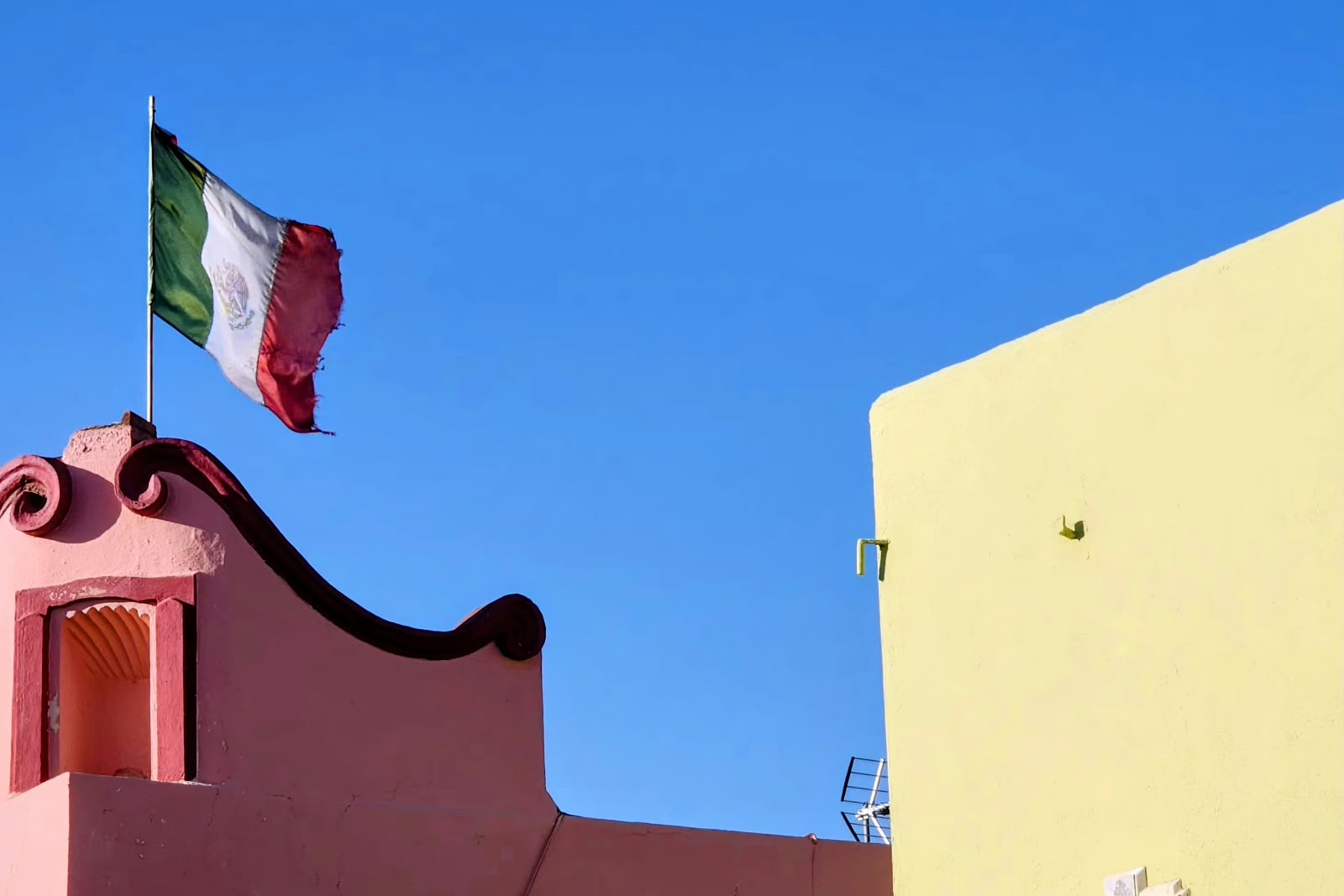 A Mexican flag flies over a pink wall and a yellow one, under a blue sky