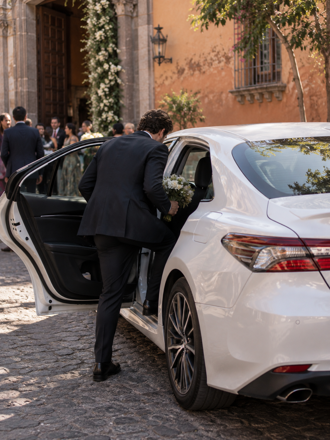 A man in a suit gets into a white car holding a bouquet of white flowers, in front of a wooden door